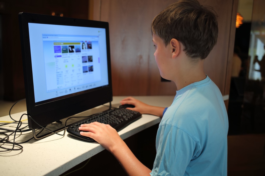 Маленький мальчик в голубой футболке сидит за компьютером в комнате один