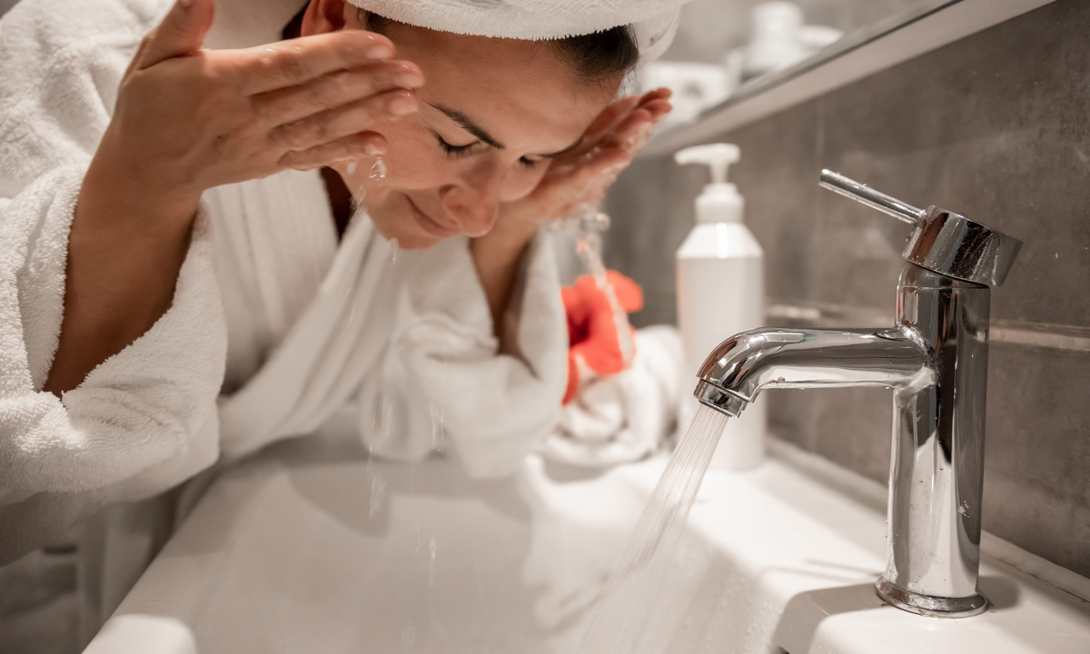 Young beautiful woman in the bathroom with a towel on her head washes her face with tap water.