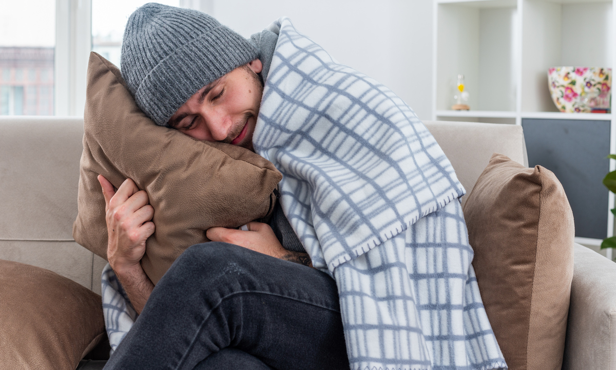 pleased young ill man wearing scarf and winter hat sitting on sofa in living room wrapped in blanket hugging pillow resting head on it with closed eyes
